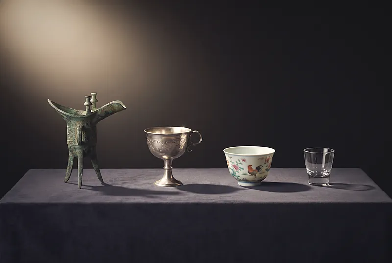 A Shang bronze jue, a Tang silver cup, a Ming porcelain cup and a modern small glass cup in a row on dark cloth