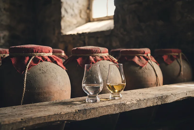 Two glasses side by side, one of new spirit and one of aged spirit, in front of old clay jars in dramatic light