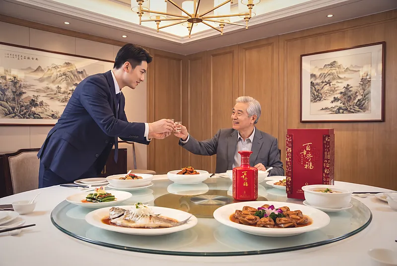 A guest at a formal Chinese business banquet raising a small cup with both hands toward an elder, with Bainian Qifu 8 on the table