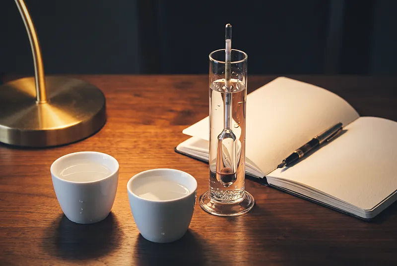 Two small cups of clear baijiu on a dark table beside an old hydrometer and a notebook
