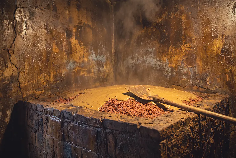 Close-up of dark, aged yellow-clay fermentation pit walls in a distillery, the grain cap sealed with mud, under warm light