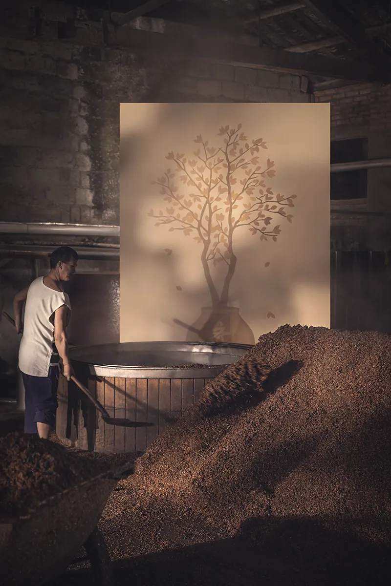 A distiller loading grain into the wooden still; the painted tree behind him is shedding its leaves