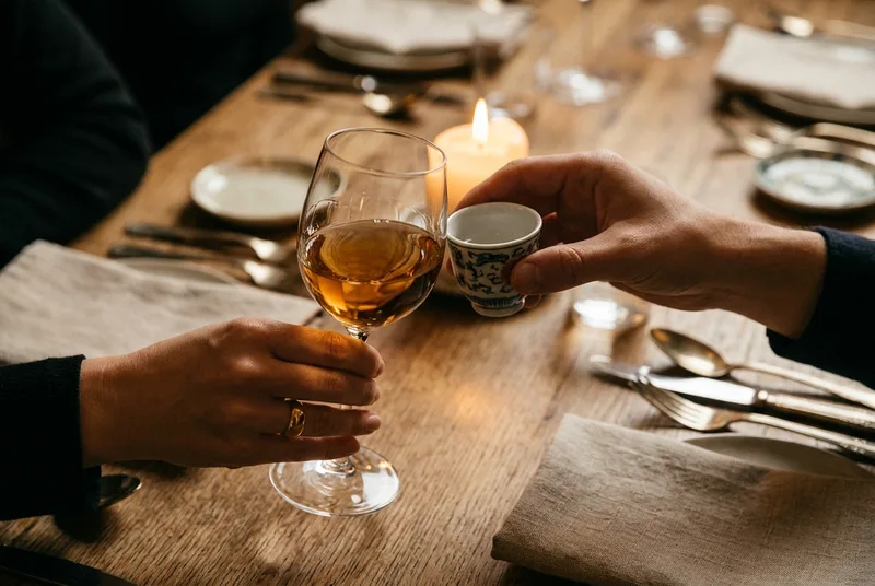 A close-up of a wine glass and a baijiu cup clinking in a toast