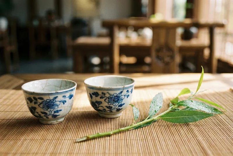 A pair of antique ceramic baijiu cups next to a snapped green willow branch on a bamboo mat